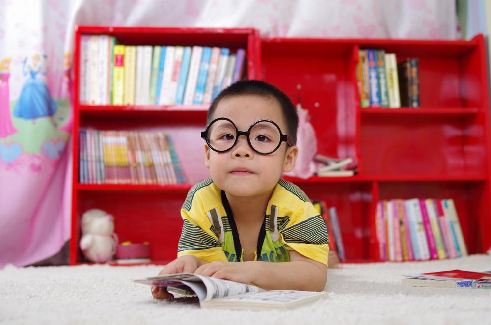 child with glasses sitting in front of bookshelf reading a book