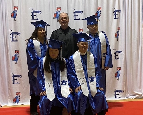 students and teacher in caps and gowns standing in front of photo backdrop of school mascot