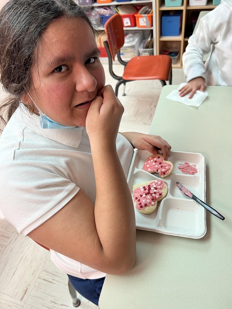 student smiling at the camera as she decorates heart shaped cookies with pink frosting and sprinkles